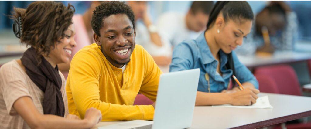  A group of diverse students in a classroom, smiling and working together. A young man in a yellow shirt is using a laptop, while others take notes.