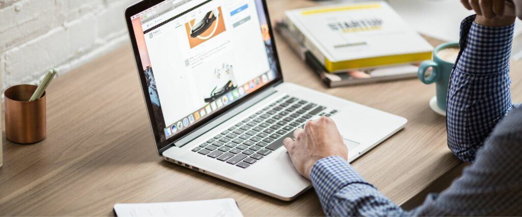 A person in a checkered shirt working on a laptop at a wooden desk, with a coffee mug, notebooks, and a pen holder nearby.